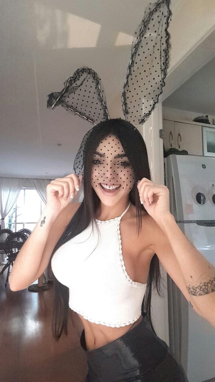 Nyvi em pose provocante segurando um controle de videogame com lingerie preta colada no corpo