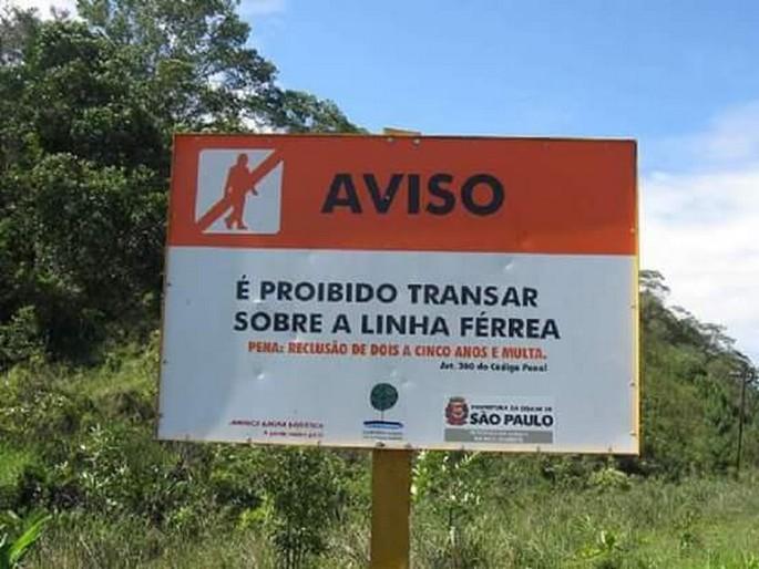 Placa de “proibido transar sobre a linha férrea” no meio do mato com céu azul e muito deboche natural